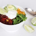 Bowl of Southwest Chili with cheddar cheese, cilantro, lime wedges, avocado, tomato, beans, corn and sour cream