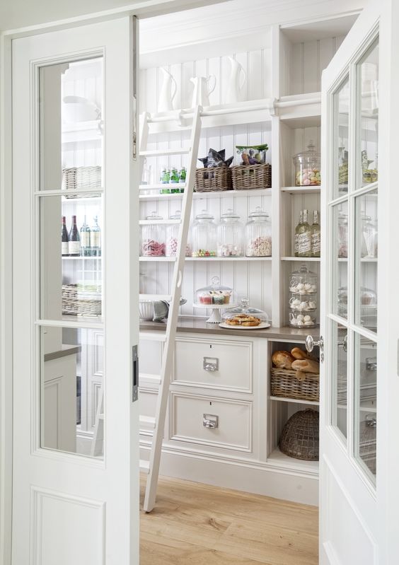 White butler pantry with glass door and ladder with apothecary jars and panelled back wall and open shelving