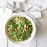 Creamy avocado pesto pasta salad in a white bowl with a spoon on a white wood table
