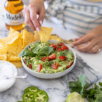 Bowl of creamy guacamole topped with chopped cilantro, diced tomatoes, onions, and lime wedges, served with tortilla chips on the side.