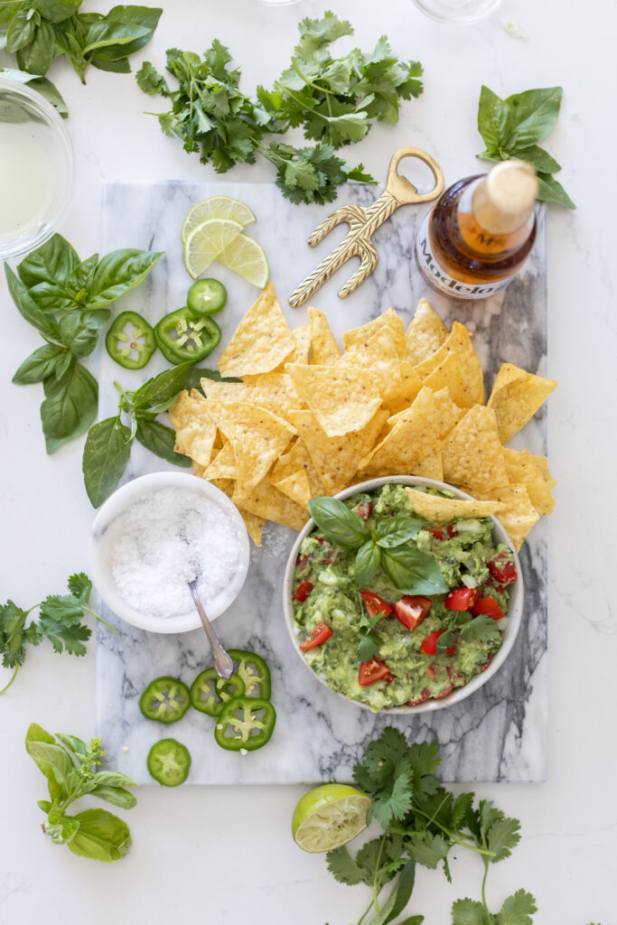 Bowl of creamy guacamole topped with chopped cilantro, diced tomatoes, onions, and lime wedges, served with tortilla chips on the side.