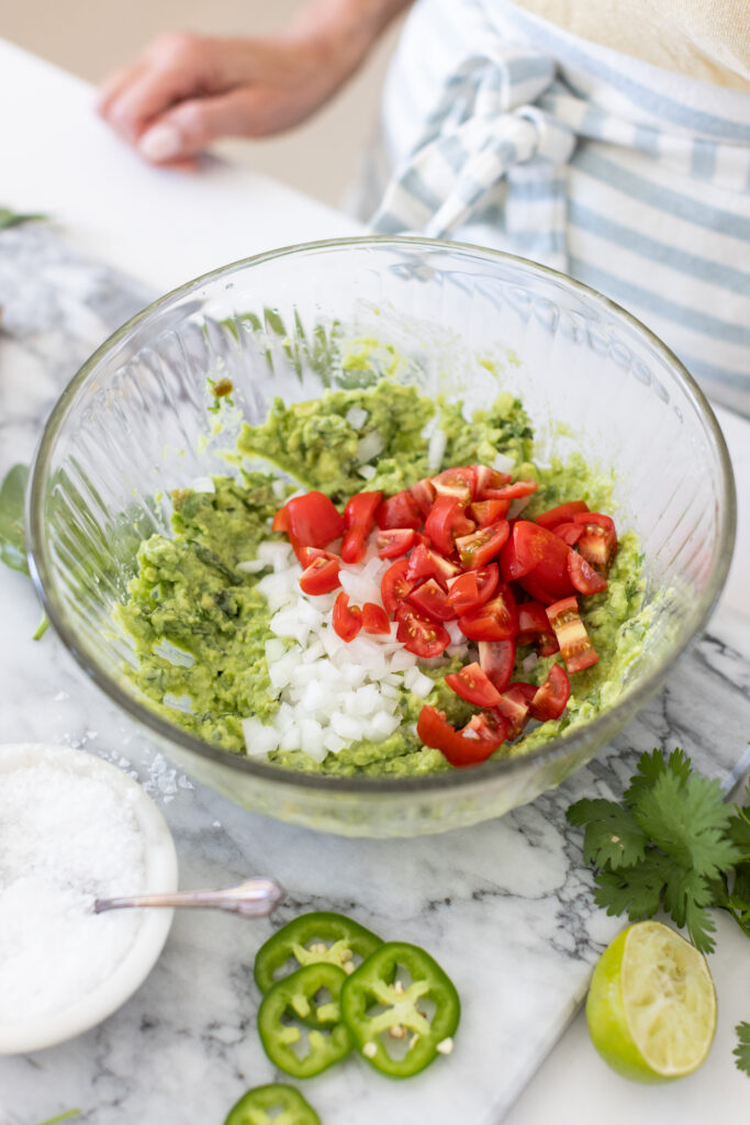 Bowl of creamy guacamole topped with chopped cilantro, diced tomatoes, onions, and lime wedges, served with tortilla chips on the side.