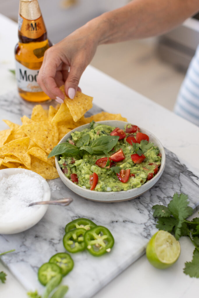 Bowl of creamy guacamole topped with chopped cilantro, diced tomatoes, onions, and lime wedges, served with tortilla chips on the side.