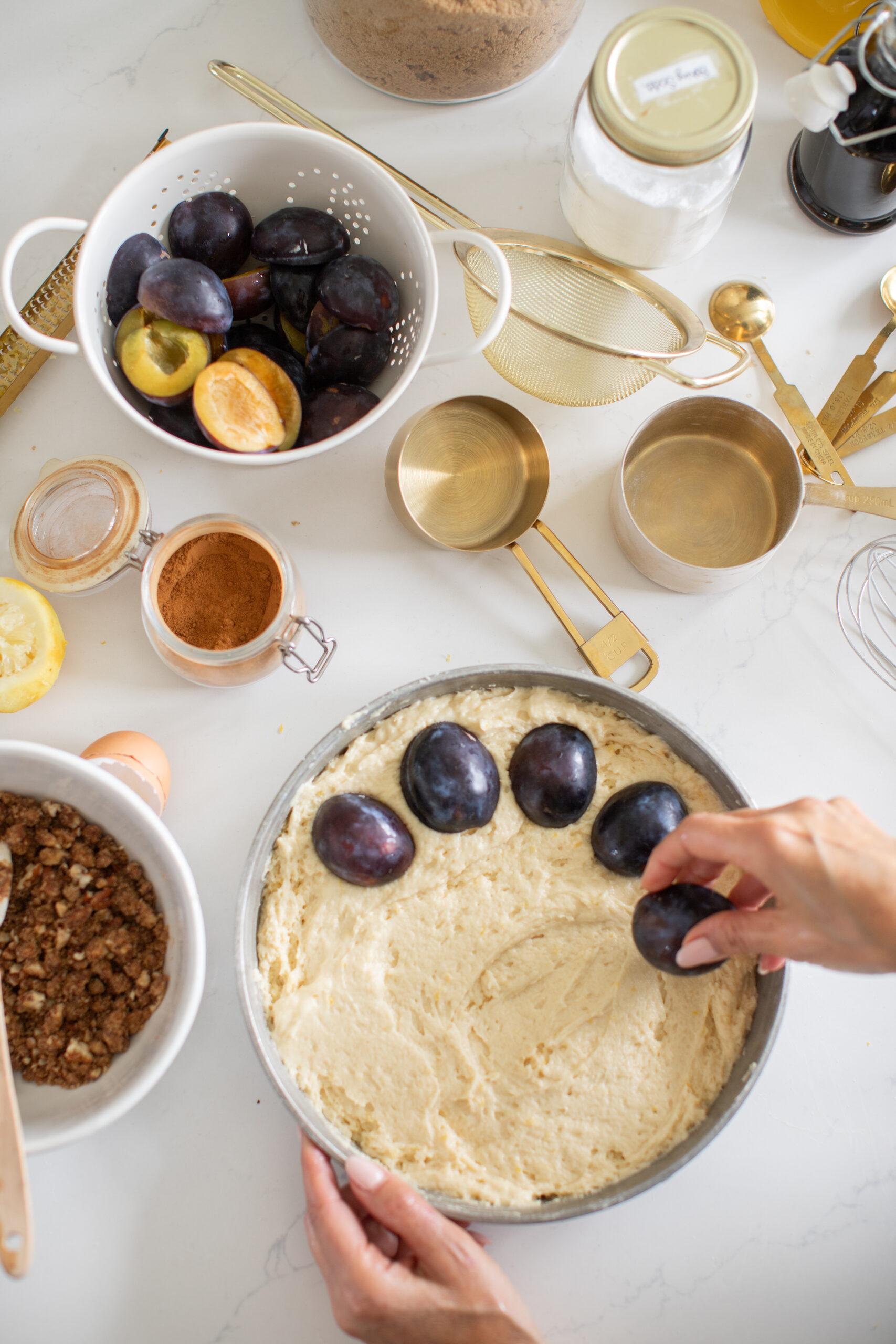 Top view of halved plums being arranged in cake batter with streusel and spices nearby.