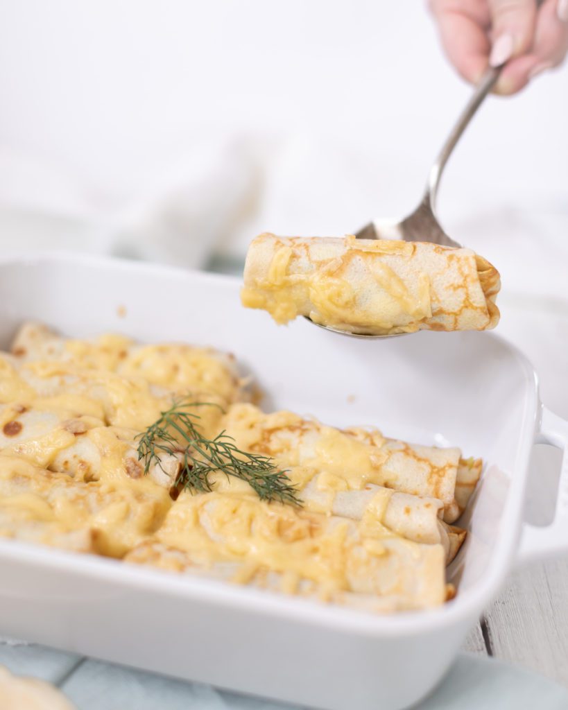 hand scooping out a piece of crepe casserole in a white baking dish topped with fresh dill and cheese