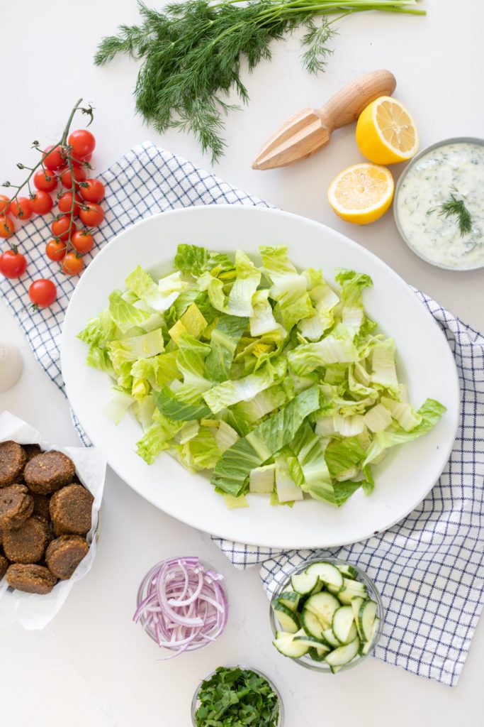 lettuce in a large bowl with falafel, cucumbers, tomatoes, dill, lemons 