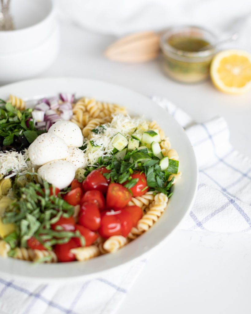  Italian Pasta Salad in a white bowl topped with tomato, mozzarella, cucumber, fresh herbs, cheese, and red onion