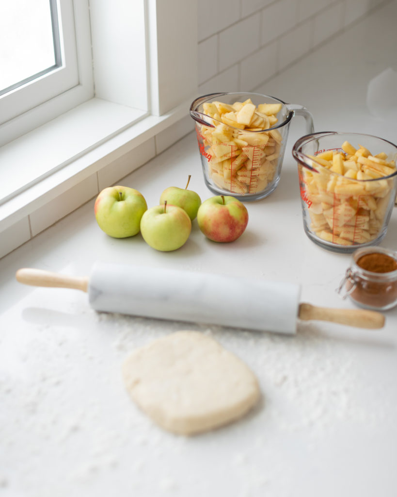The perfect pie dough on a counter with a rolling pin and jars of sliced apples