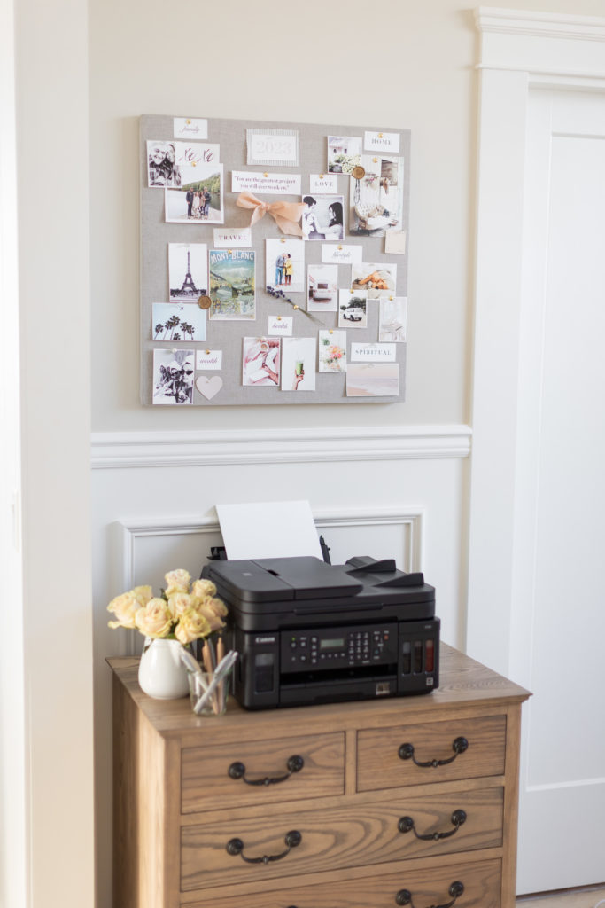 a DIY Vision Board hanging on the wall above a black Canon printer on a desk with a vase of white roses