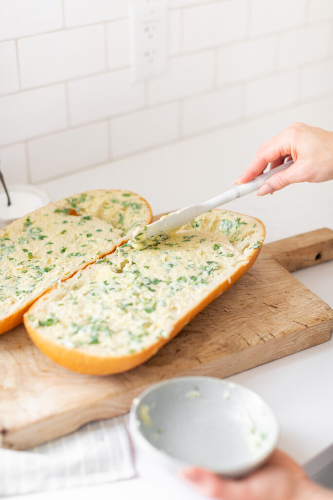 Two halves of a French Loaf on a wooden cutting board and a hand is spreading garlic butter on the bread to make it into Garlic Bread. 