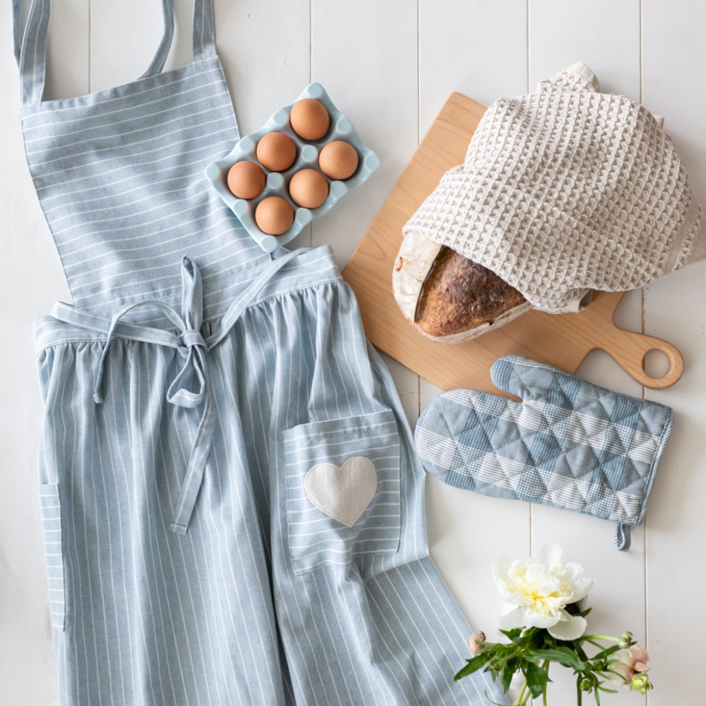 A blue pinstripe apron and oven mitts from the Fraiche Food, Fuller Hearts Collection by The Jilly Box, next to a carton of eggs and a fresh load of bread. 