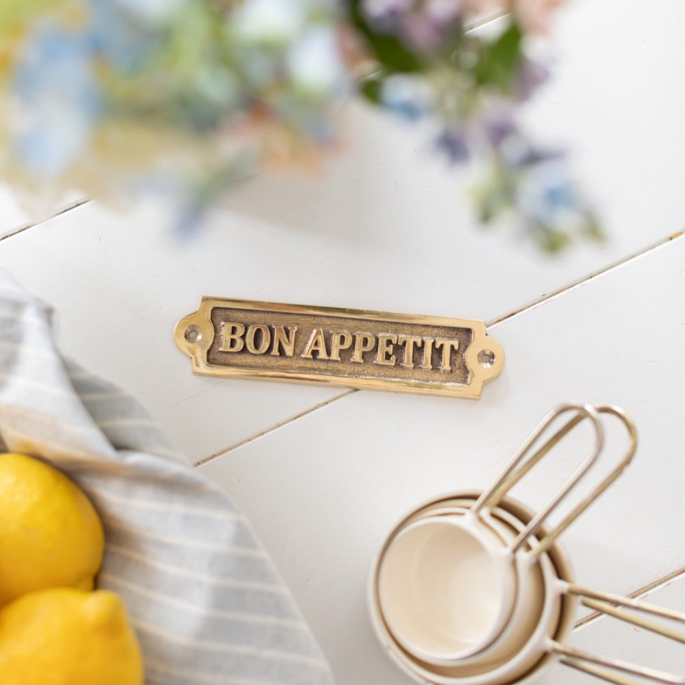 brass bon appettit wall sign on a white table surrounded by measuring cups, flowers and lemons