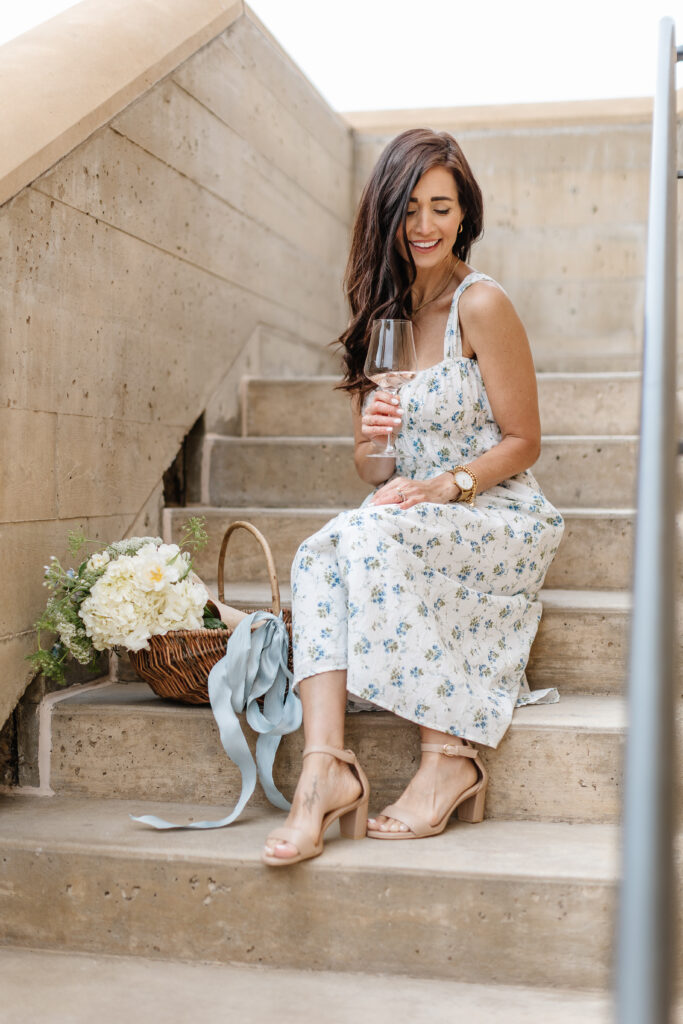 Tori sitting on steps in her blue and white floral dress with a glass of wine from The Fraîche x PRIV Summer Collection