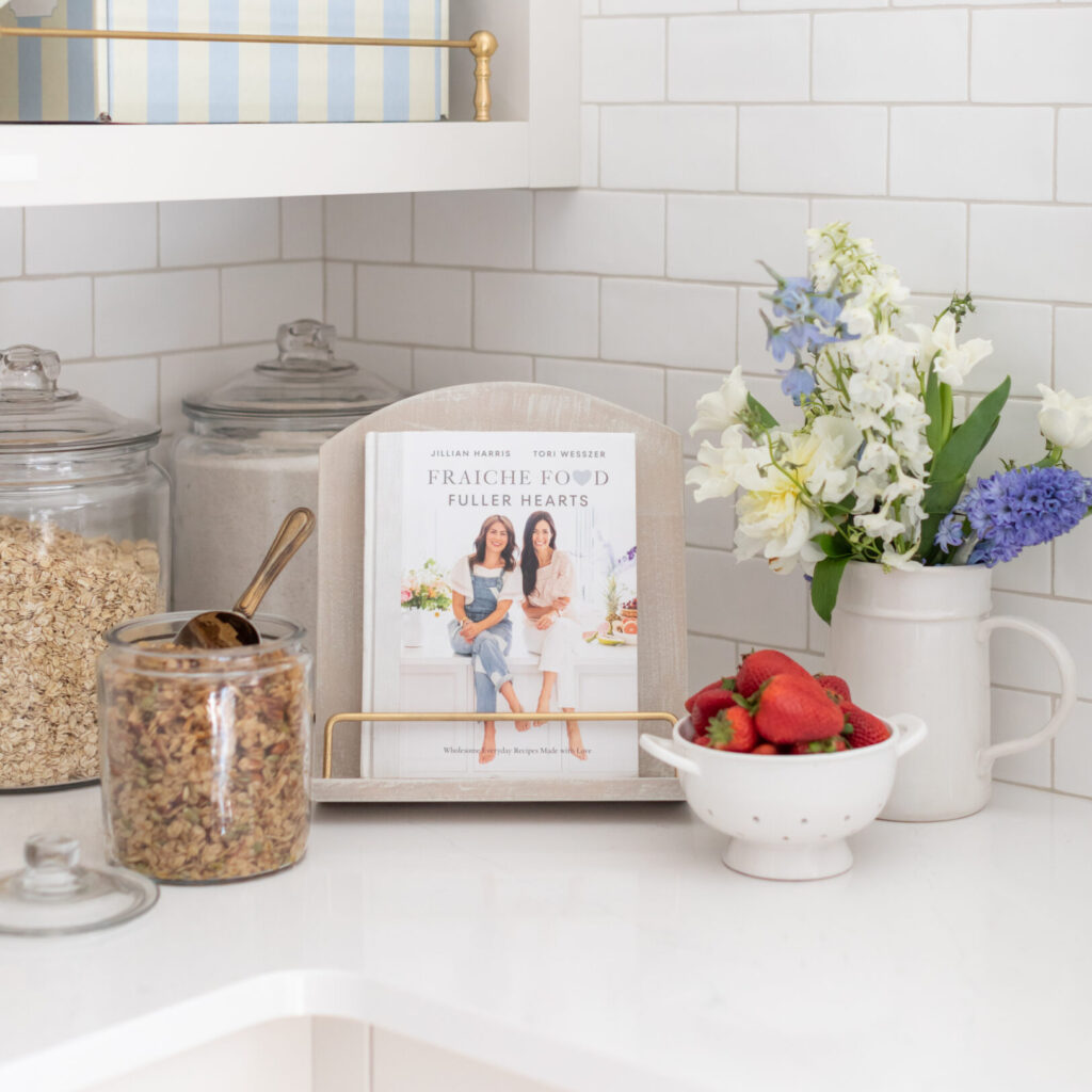 The Fraiche Food, Fuller Hearts Cookbook on a wooden cookbook stand, next to a jar of granola, bowl of berries and a vase of flowers.