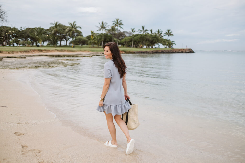 Tori on the beach in Hawaii walking with sandals in the water in a blue tee shirt dress from The Fraîche x PRIV Summer Collection
