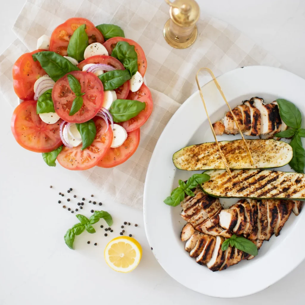 Caprese salad on a plate with tomato, basil, mozzarella and balsamic, beside a tray of grilled chicken and zucchini