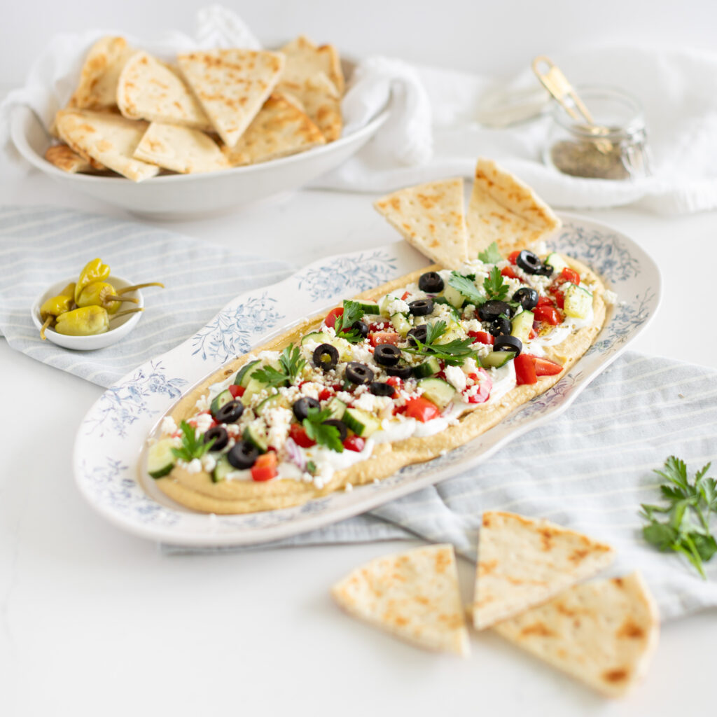 a 7-Layer Greek Dip with hummus, tzatziki, tomatoes, cucumbers, feta and olives, on a tray, served beside pita bread