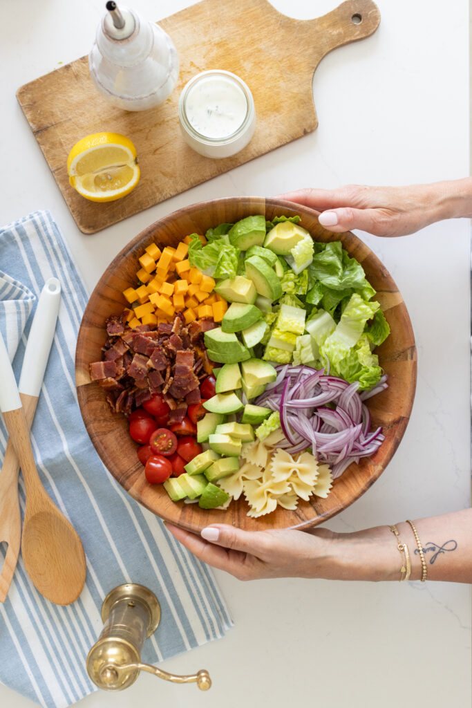 two hands holding a large bowl of BLT pasta salad with bowtie pasta, crispy bacon, creamy avocado, juicy tomatoes and cheddar cheese