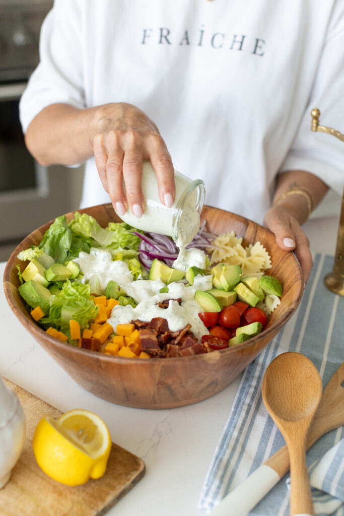 pouring ranch dressing over a bowl of BLT pasta salad with bowtie pasta, crispy bacon, creamy avocado, juicy tomatoes, cheddar cheese, and romaine lettuce.