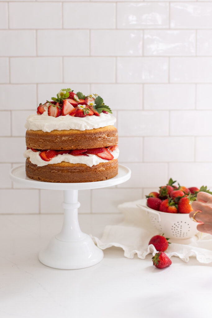 Strawberry Shortcake Cake sits on a white cake pedestal topped with strawberries, whipped cream and flowers