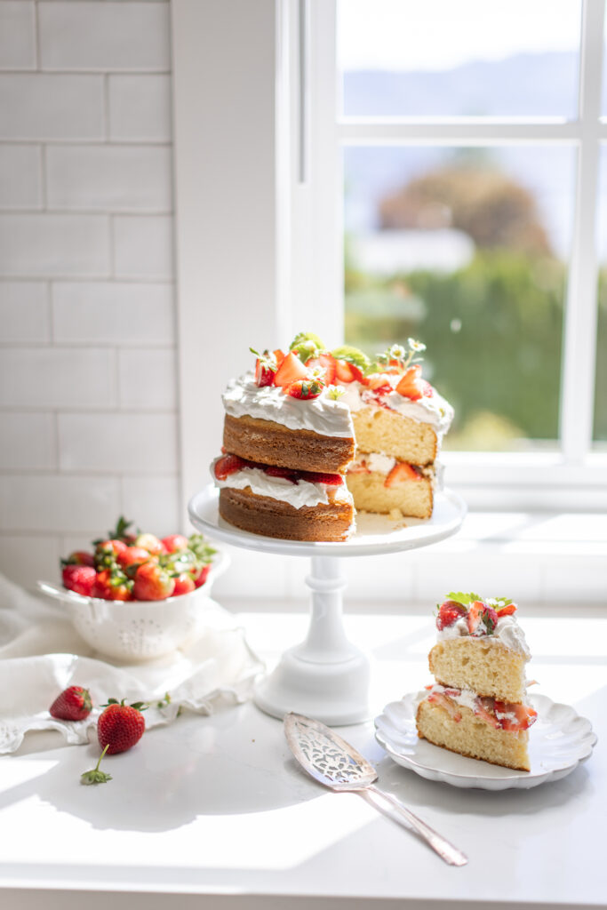 a Strawberry Shortcake Cake sits on a cake pedestal in front of an open window topped with strawberries and edible flowers