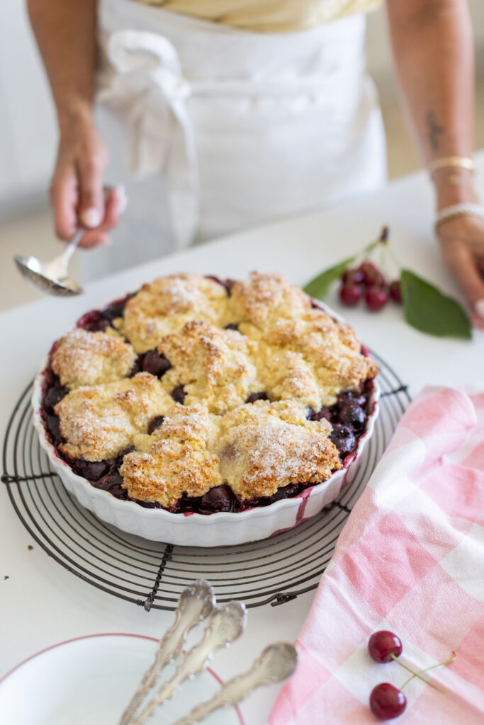 A classic cherry cobbler baked until golden, with bubbling sweet cherries and a rustic biscuit topping in a white baking dish.