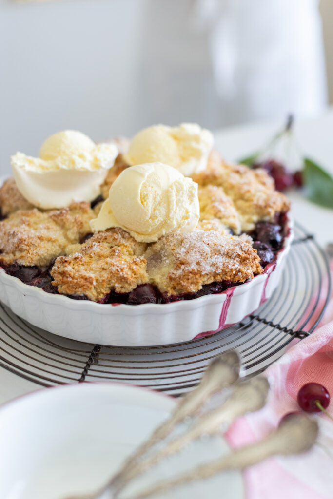 A classic cherry cobbler baked until golden, with bubbling sweet cherries and a rustic biscuit topping in a white baking dish.