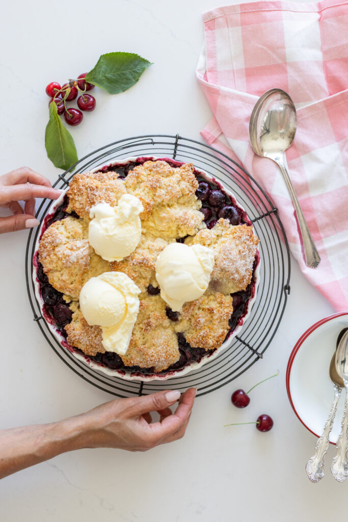A classic cherry cobbler baked until golden, with bubbling sweet cherries and a rustic biscuit topping with vanilla ice cream in a white baking dish.