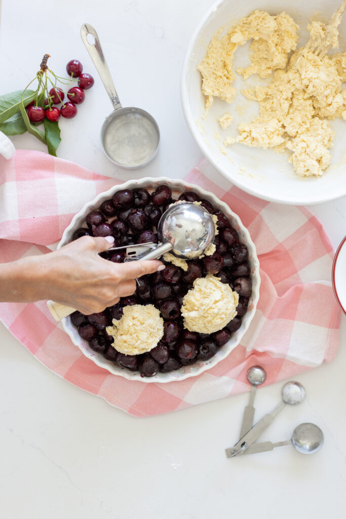 Unbaked classic cherry cobbler in a baking dish, with juicy red cherry filling and biscuit dough being spooned or placed on top before baking.