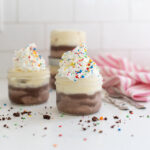 Close-up of a finished Dairy Queen Ice Cream Cake Jar, layered with chocolate and vanilla ice cream, fudge sauce, and a crunchy cookie layer, topped with whipped cream, sprinkles in a clear glass jar.