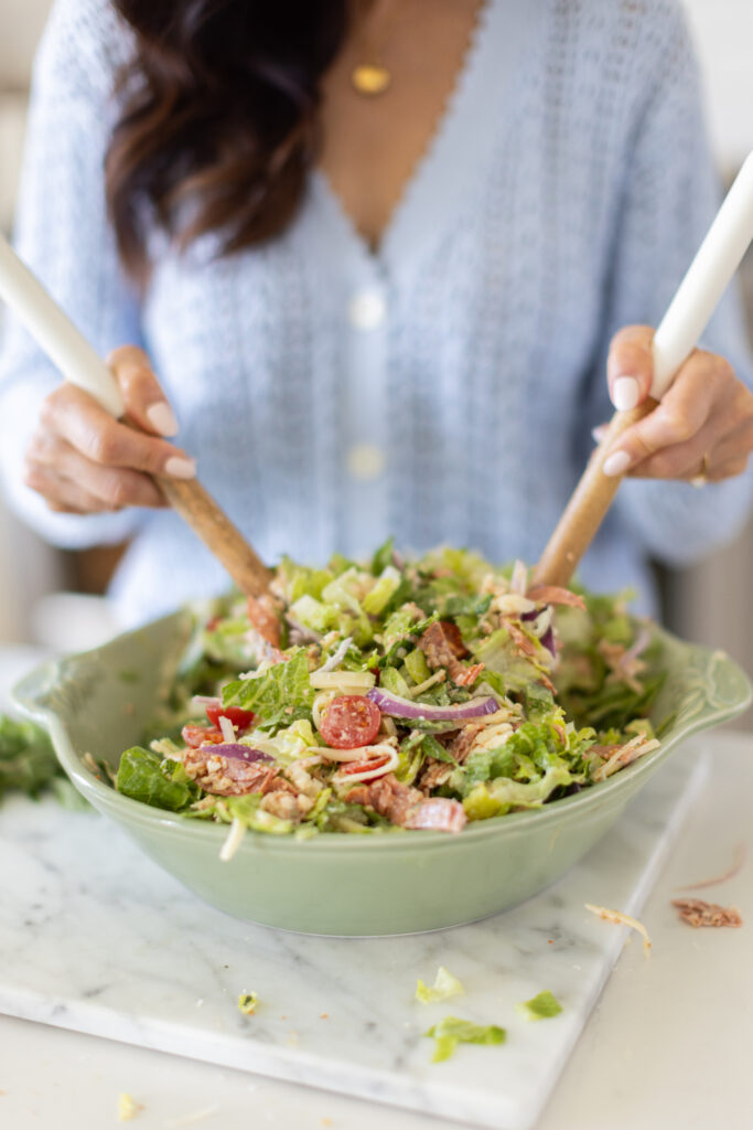 Tori in a blue shirt standing in a bright kitchen, tossing Pizza Ranch Salad in a large green bowl with wooden salad servers.