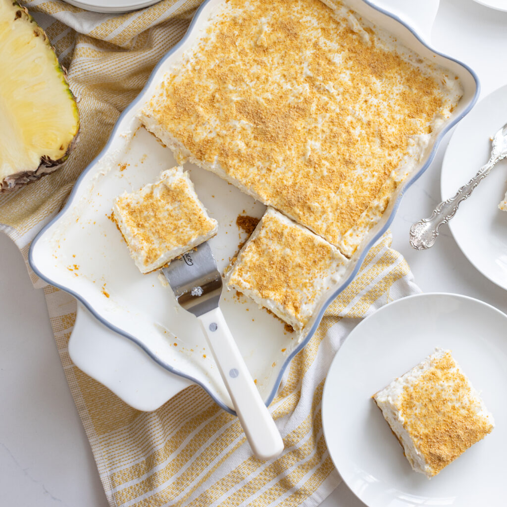 No-bake Pineapple Delight dessert with graham cracker crust, cream cheese filling, in baking dish with serving plates on the side