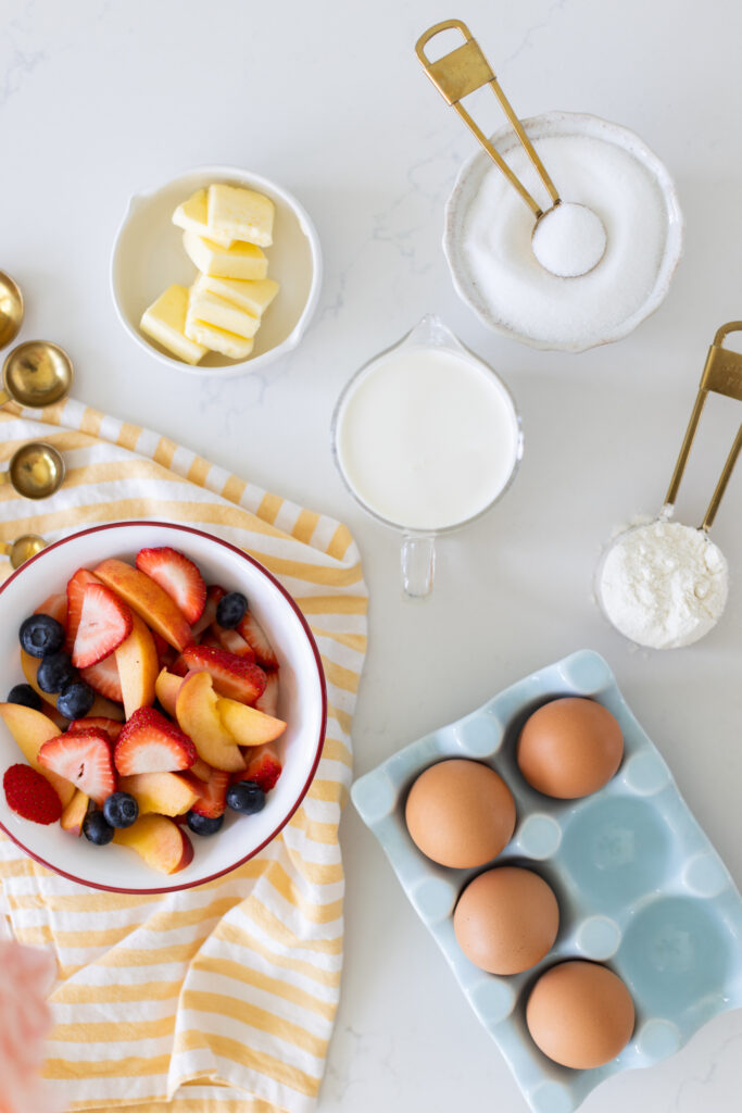 Ingredients for sheet pan crepes displayed on a white counter, including fresh strawberries, blueberries, sliced peaches, butter, milk, sugar, flour, and eggs.