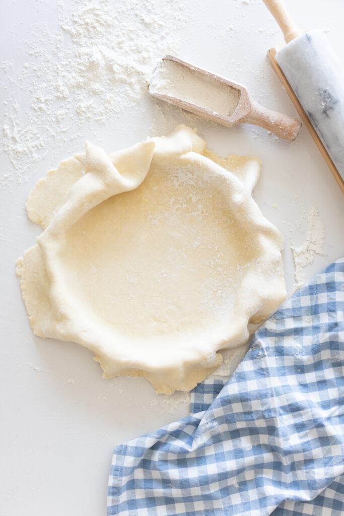 homemade pie crust in a white dish, with flour, rolling pin, and blue gingham towel on the counter.