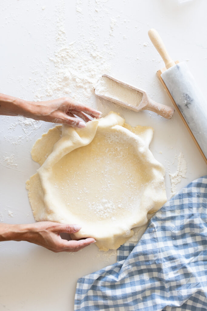 Hands shaping homemade pie crust in a white dish, with flour, rolling pin, and blue gingham towel on the counter.