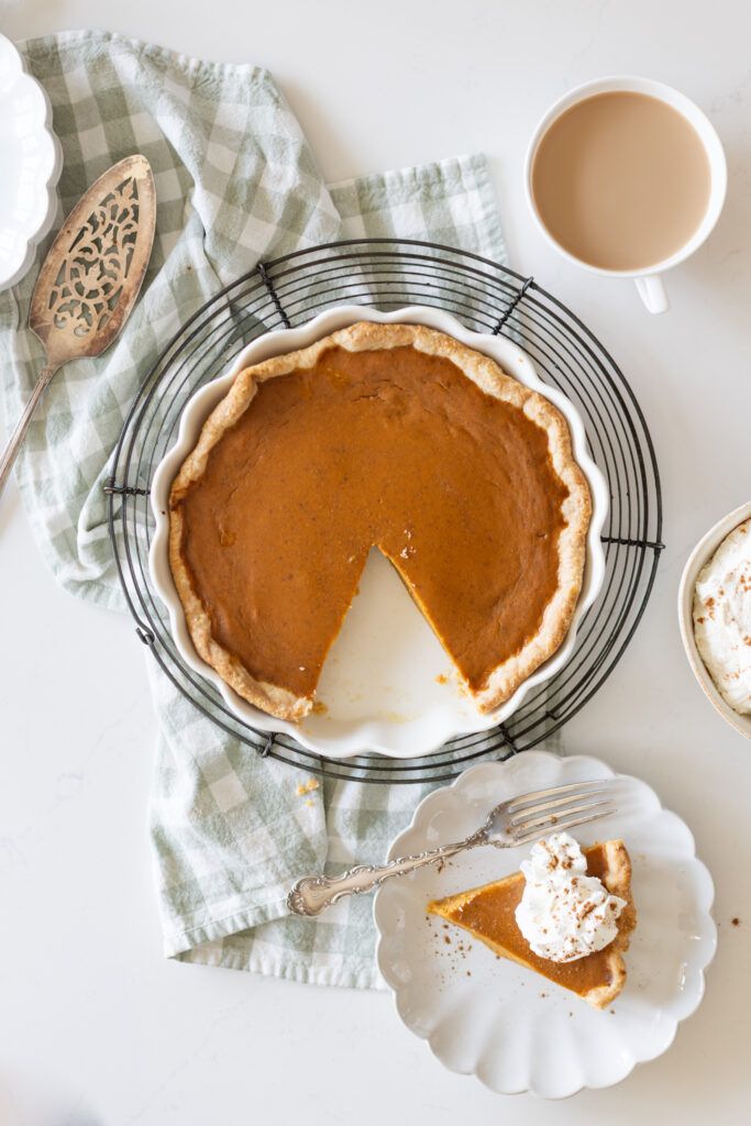 Best pumpkin pie in a white pie dish with one slice removed, slice on a plate topped with Chantilly Cream, served with coffee on a fall table.