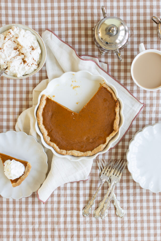 Pumpkin pie in a white pie dish with one slice removed, slice on a plate topped with Chantilly Cream, served with coffee on a fall table.